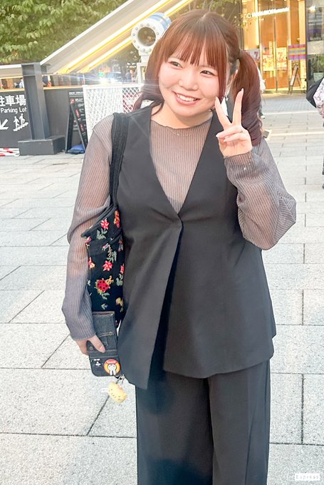 A woman with reddish-brown pigtails smiling and making a peace sign on a city street, wearing a sheer gray blouse over a black top with a floral tote bag.