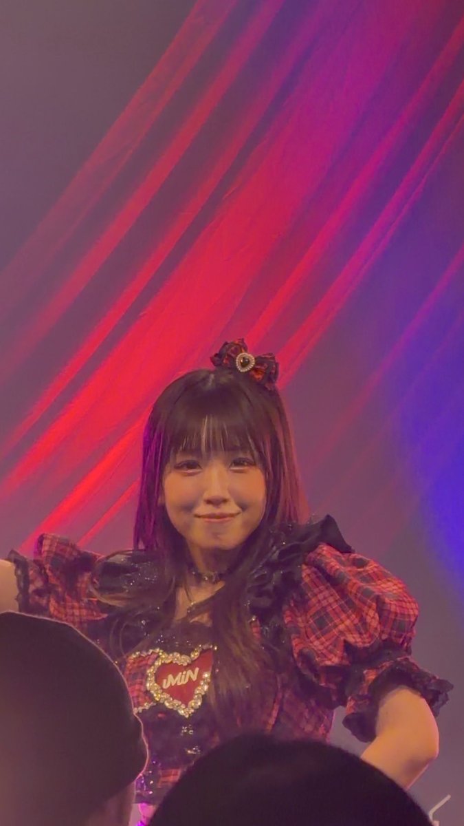 A performer in a red and black plaid outfit with a heart badge smiling under red stage lighting.