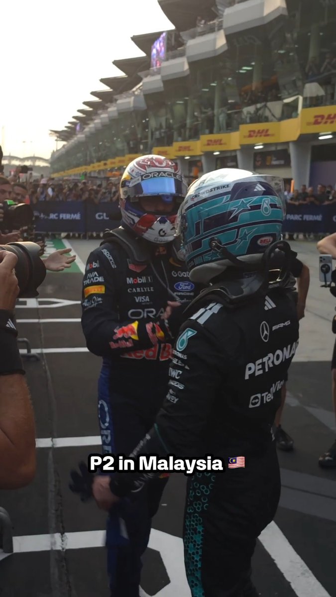 Two Formula 1 drivers embrace in parc fermé, with one wearing a Red Bull Oracle racing suit and the other in Mercedes Petronas gear. Visible text includes sponsor logos like "Oracle," "Mobil 1," "Petronas," "IWC," and "Bell," along with caption text reading "P2 in Malaysia" with a Malaysian flag.