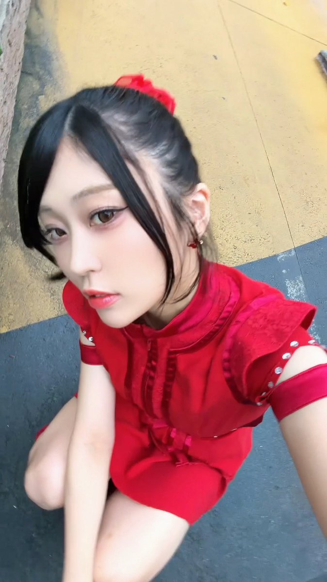 A young woman in a short red dress with a red bow in her hair looks up at the camera while crouching on a sidewalk next to a yellow wall.