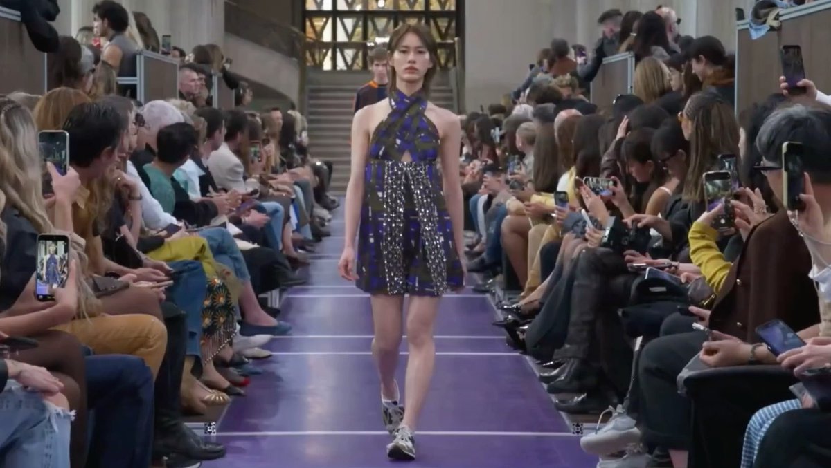 A model walks down a purple runway at the Miu Miu SS27 fashion show, wearing a navy blue patterned halter dress with a sparkling silver waist detail, while audiences on both sides capture the moment on their phones. The image aligns with the Thai caption celebrating "เอมี่" (Emi) representing on the prestigious international runway at the Miu Miu Spring/Summer 2027 showcase.