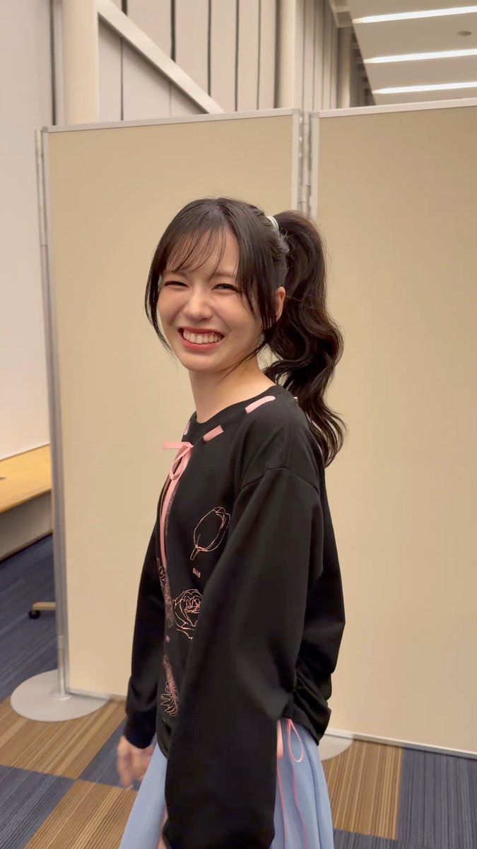 A young woman with a long ponytail smiles brightly in what appears to be an indoor studio or backstage area, wearing a black long-sleeved top with pink ribbon detailing and floral embroidery. The hashtag "#Girls2" in the post likely refers to the Japanese idol group Girls², suggesting this is a photo from their fan send-off event ("お見送り会"), capturing a candid moment with one of the members.