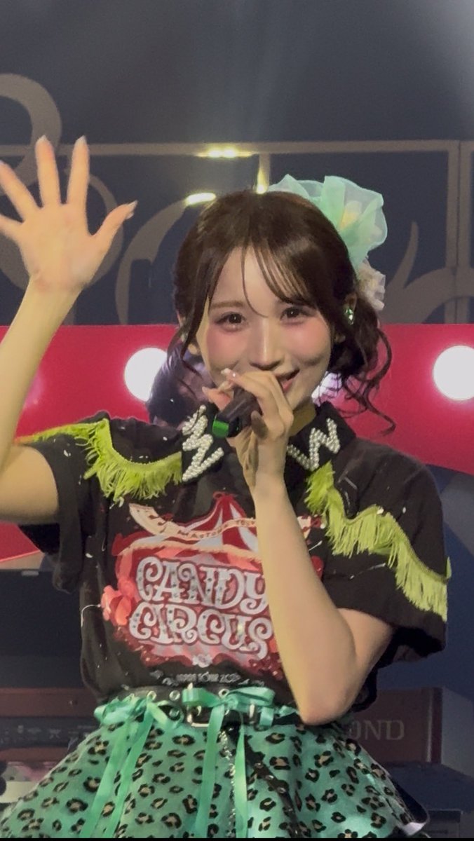 A young woman performs on stage wearing a black 'CANDY CIRCUS' shirt with green fringe and a matching green and black polka-dot skirt, holding a microphone and waving.