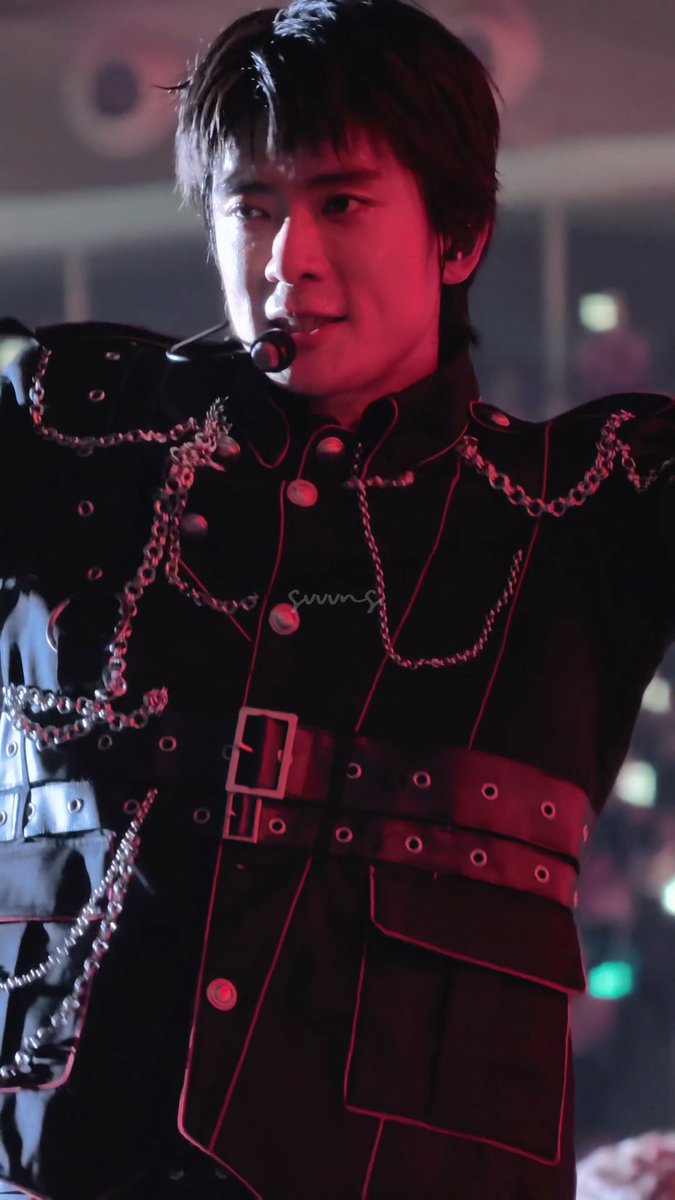 A young man, likely a performer, wearing an elaborate black military-style outfit adorned with silver chains and a belt, with a headset microphone, illuminated by red stage lighting.