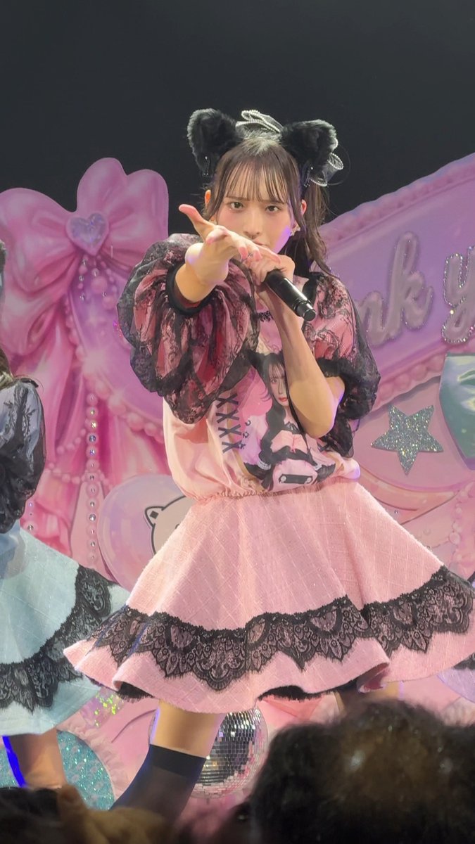 A young woman performs on stage in a pink and black gothic-style dress, pointing toward the camera with a microphone in the other hand.