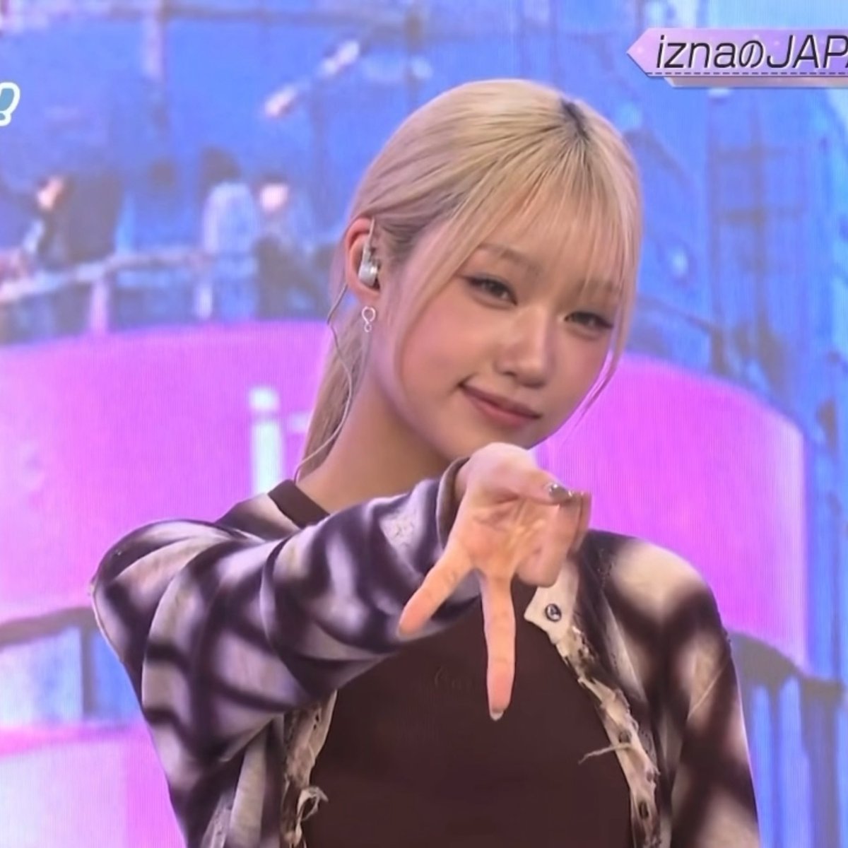 A blonde female performer on a stage with pink and blue lighting, wearing a patterned jacket, extending her hand toward the camera with a soft smile. Text 'iznaのJAP' is visible in the upper right corner.