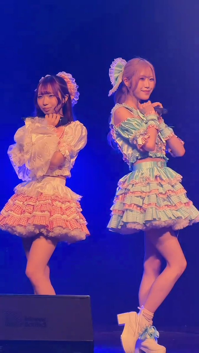 Two female performers in colorful, frilly pastel idol outfits pose on a dimly lit blue stage, one in a pink and yellow dress and the other in a green and yellow dress.