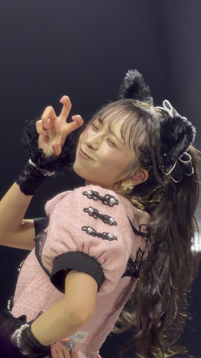 A young woman with dark wavy hair styled in a high ponytail with bows performs on stage in a pink ruffled dress with black trim, posing with her hand near her face.