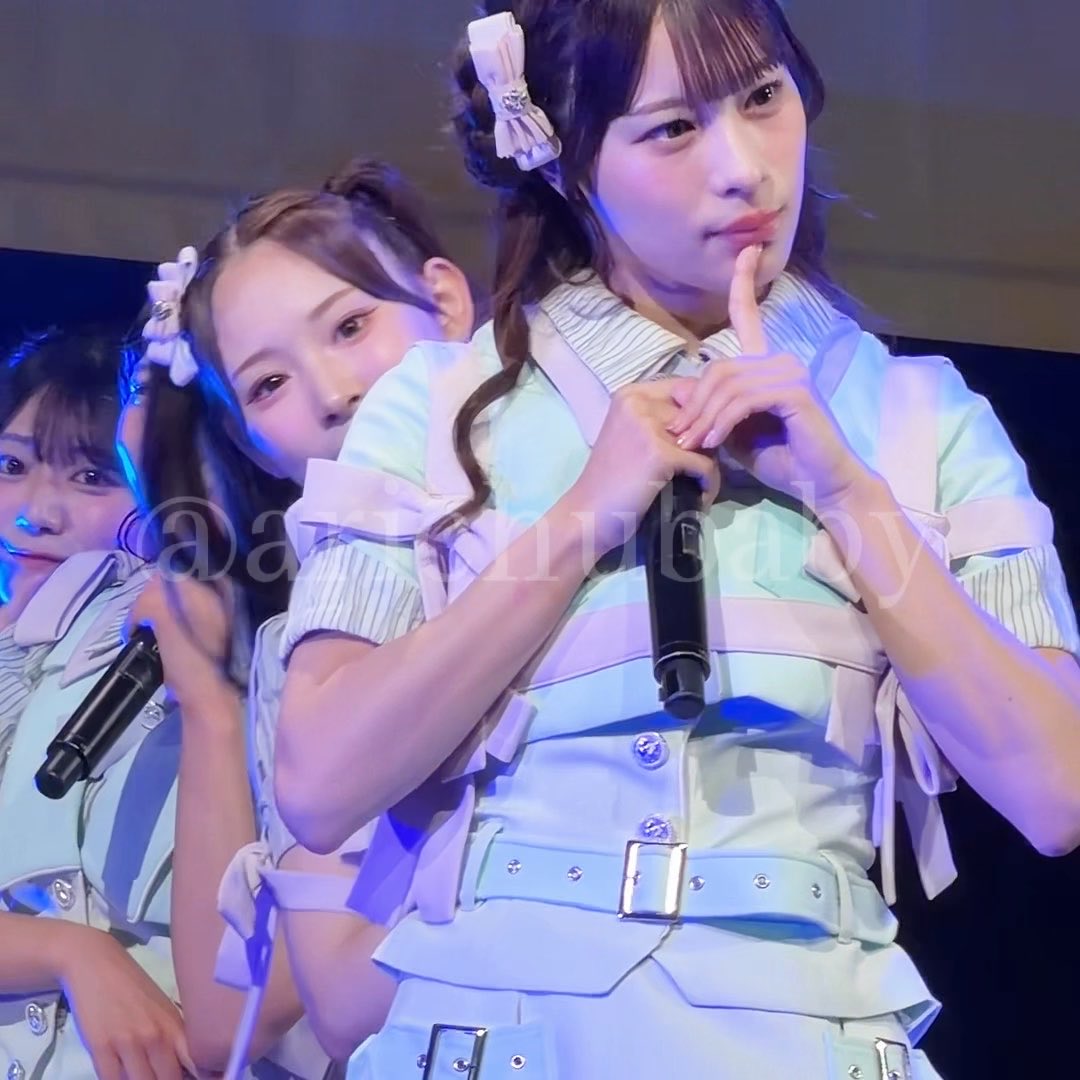 Three young women in matching light green and white idol outfits perform on stage, with the front girl making a heart shape with her hands near her face.