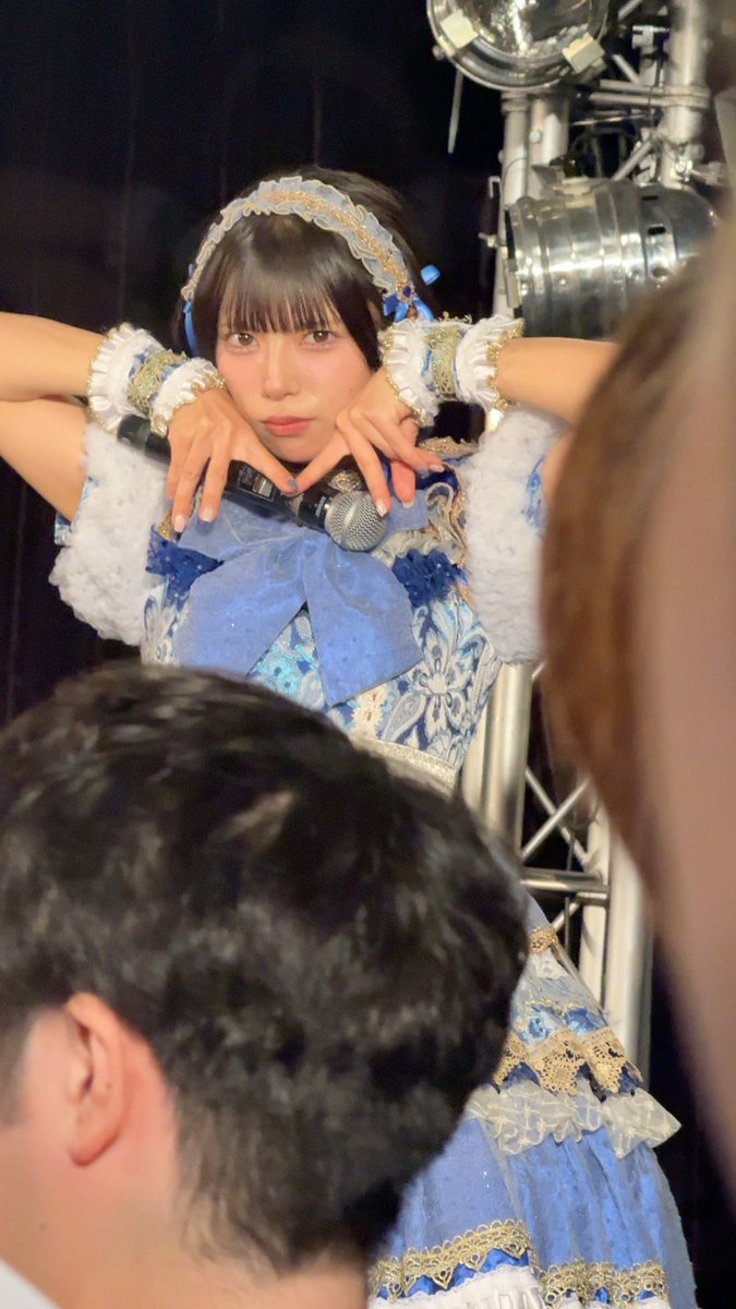 A young woman wearing an elaborate blue and white frilled idol costume with a matching headband holds a microphone while posing backstage.