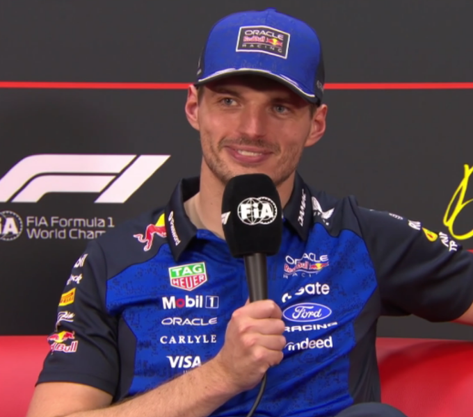 Max Verstappen, wearing his Red Bull Racing cap and team shirt featuring sponsors like Oracle, TAG Heuer, Mobil, and Visa, smiles while speaking into an FIA-branded microphone during a press conference, tying into the joke about Oracle's "fast connection" enabling quick software updates.