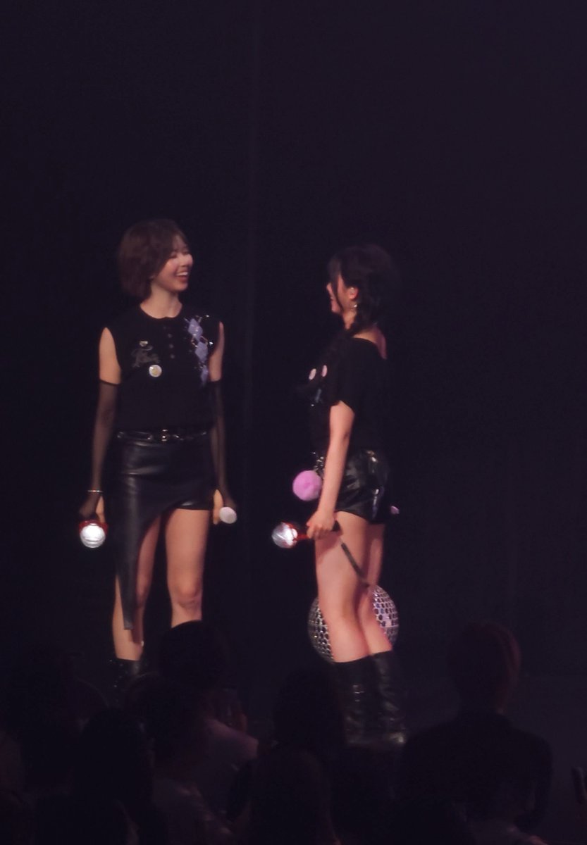 Two female performers in black outfits hold microphones on a dimly lit concert stage.