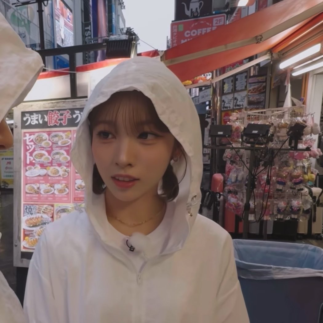 A young woman wearing a white hooded raincoat stands in front of a Japanese street food stall with menus visible in the background.