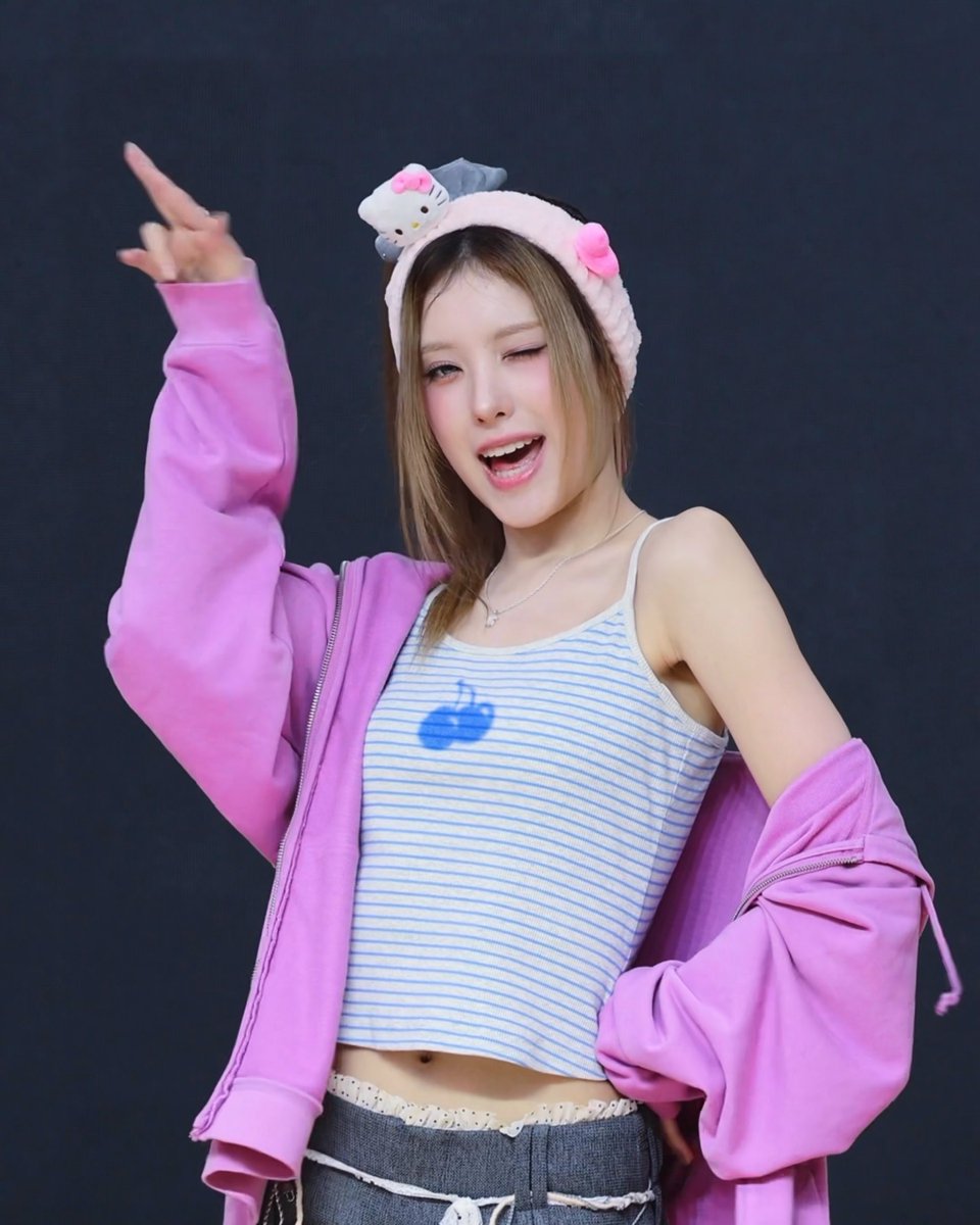 A young woman poses energetically in a pink zip-up jacket over a blue striped crop top, wearing a Hello Kitty headband and winking.