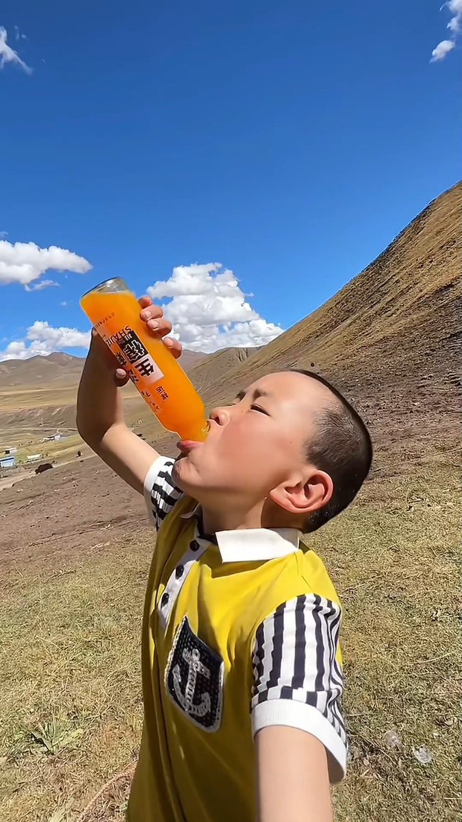 A young boy in a yellow and black striped shirt drinking an orange soda from a glass bottle in a mountainous landscape under a blue sky.