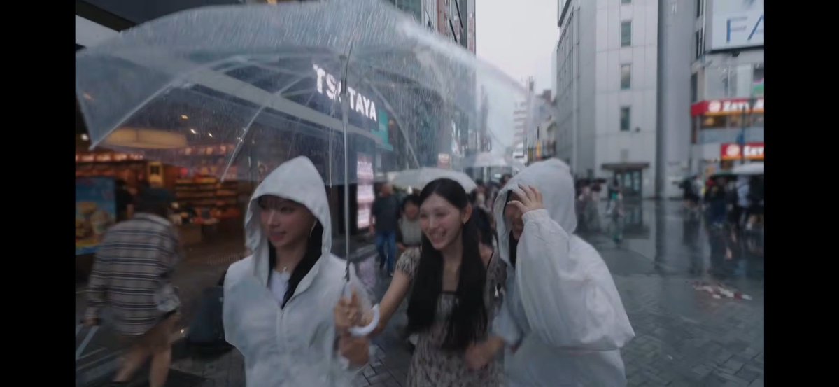 Three young women in rain ponchos share a transparent umbrella while walking through a rainy shopping street in Japan, with a 'TSUTAYA' store visible in the background.