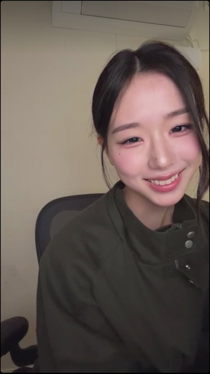 Selfie-style portrait of a smiling young woman wearing a dark green jacket, sitting in a chair.
