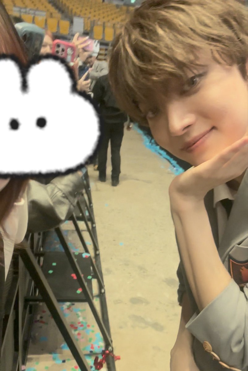 A young man with light brown hair poses for a close-up photo at what appears to be a fan event, resting his chin on his hand while a pixelated heart sticker covers the face of another person in the foreground. Confetti scattered across the floor and a barrier separating fans suggest this was a post-show meet-and-greet, aligning with the post about Yiju (의주) kindly reaching out to a fan whose hand was blocked by staff.