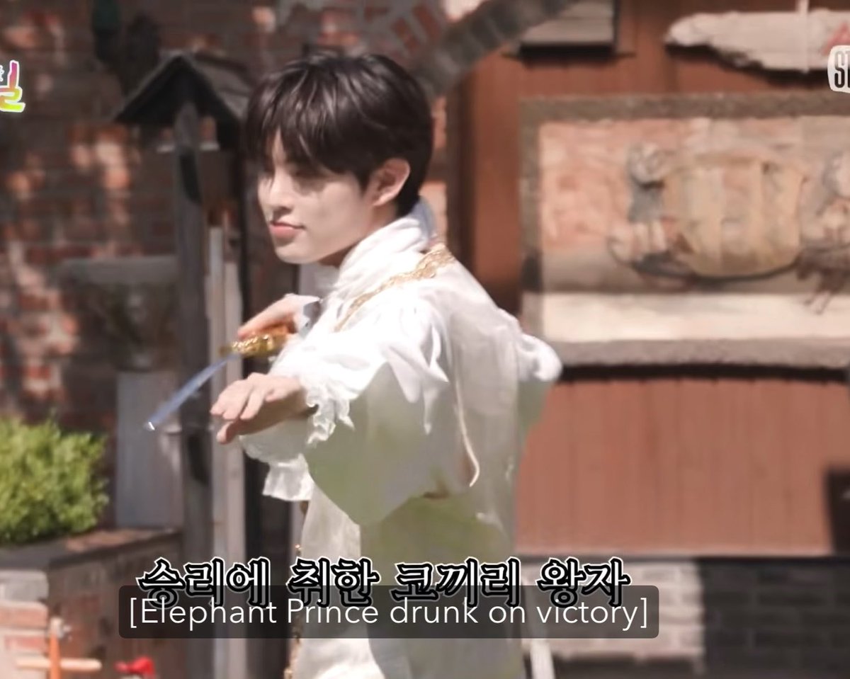 A young man with dark hair in a white ruffled shirt holding a sword-like object, with the Korean and English subtitle "승리에 취한 코끼리 왕자 [Elephant Prince drunk on victory]."