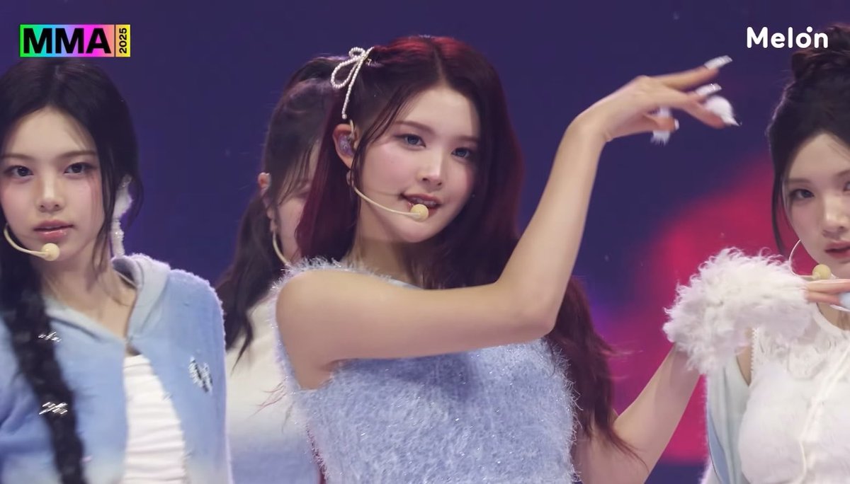 Three K-pop performers are on stage at MMA 2025 (logo visible top left, Melon logo top right), with the central woman in a fuzzy light blue top gesturing with her hand raised.