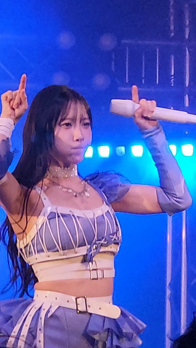 A young woman with long dark hair performs on stage in a blue and white corset-style outfit, holding a white microphone and pointing upward with blue stage lighting behind her.
