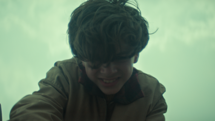 A young boy with tousled dark hair smiles downward against a hazy, overcast sky, his face partially shadowed by his locks. The eerie green-tinted atmosphere and enigmatic expression evoke the mysterious "something in the water" teased by the upcoming premiere of BELOW on October 8.