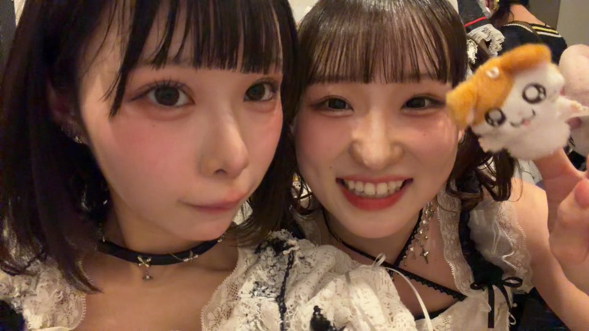 Two women with matching dark hair and bangs pose together for a selfie, both wearing white lace outfits, with one holding up a small plush toy.