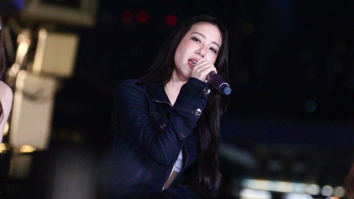 A young woman with long dark hair singing into a microphone while wearing a dark denim jacket, appearing to perform on stage at night.