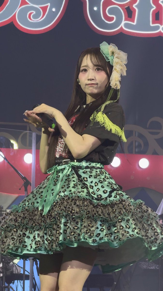 A young woman with long dark hair and a floral hair accessory performs on stage in an elaborate green and black leopard-print ruffled dress with neon yellow fringe, holding a microphone.