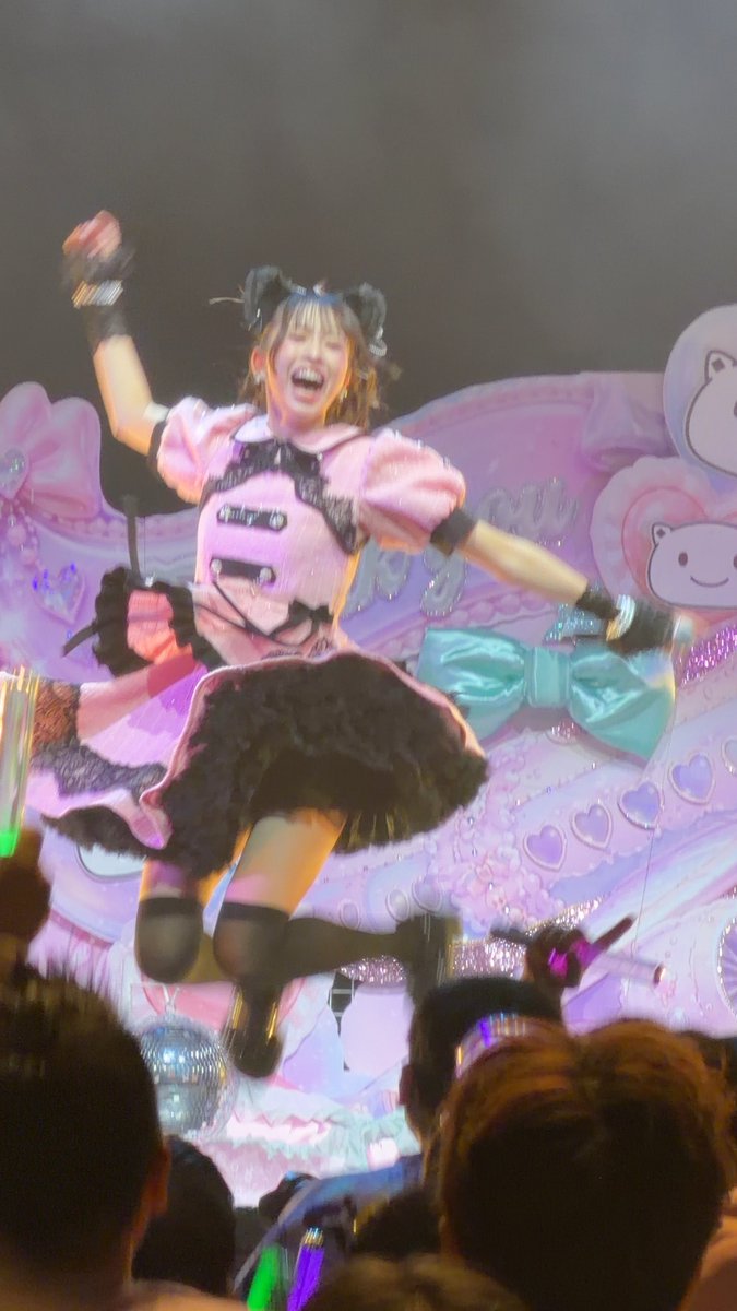 A performer in a pink and black frilly idol costume jumps mid-air on a candy-themed stage with cartoon bear decorations, smiling joyfully with her arms outstretched. The hashtags #わーすた (Wasuta) and #三品瑠香 (Mishina Ruka) confirm this is a concert moment featuring Ruka from the J-pop group Wasuta, captured by a fan referencing a moment they found amusing with the caption "『ひくいな』" (something like "that's pretty cheap/amateurish" or "that's lame").