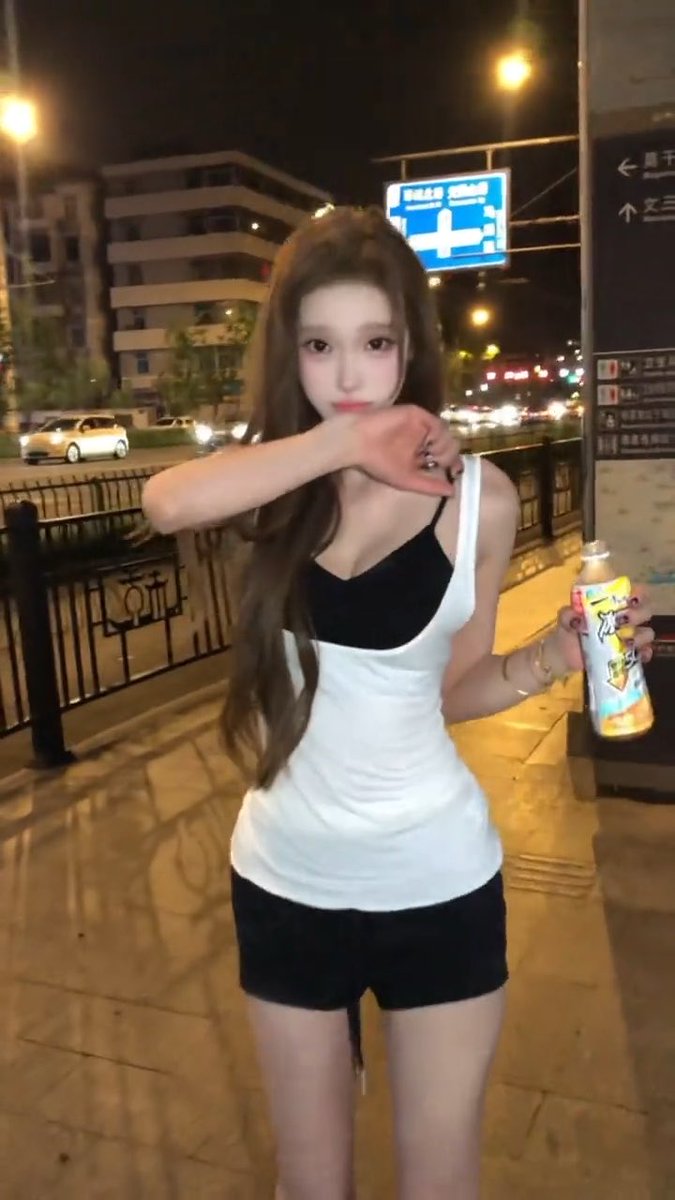 A young woman in a black and white outfit poses on a city street at night holding a colorful spray bottle, with Chinese characters visible on directional signs behind her.