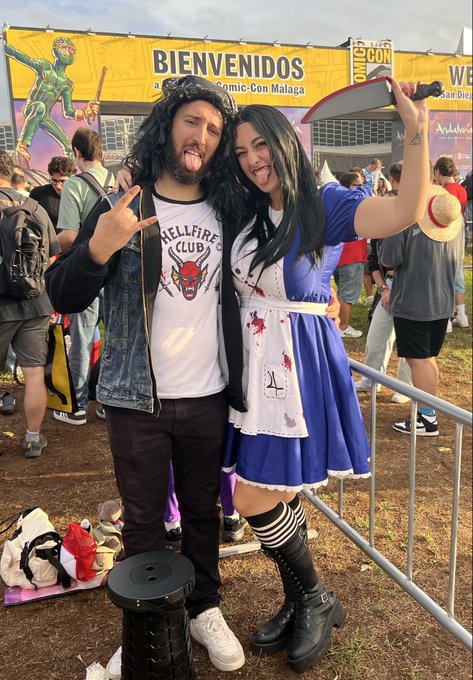 Two excited cosplayers pose at SDCC Málaga in front of a yellow "BIENVENIDOS" banner—one dressed as a rocker in a "Hellfire Club" shirt holding a prop guitar, and the other in a bloody blue Alice in Wonderland-style dress wielding a knife and straw hat—capturing the fun and fandom energy of the event where the poster celebrated getting signatures from celebrities like Elijah Wood.