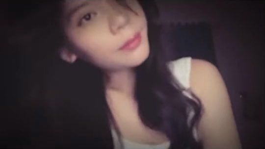 A blurry, low-light selfie-style video frame of a young woman with long dark hair wearing a white top.