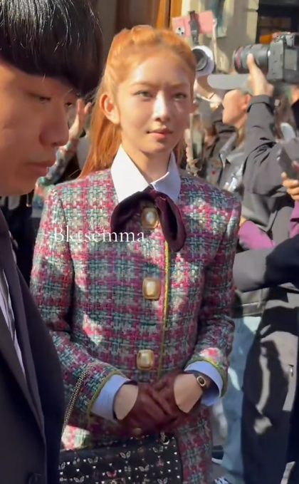 A woman with long orange hair stands outdoors in a tweed jacket with a burgundy bow tie, flanked by a man and surrounded by photographers; the watermark 'blkdstmnm9' is visible.
