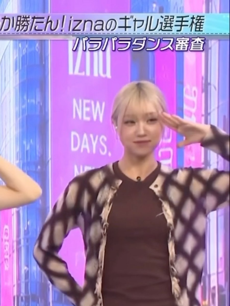 A blonde woman salutes during a Japanese TV broadcast, with on-screen text reading 'iznaのギャル選手権 パラパラダンス審査' against a backdrop featuring 'IZNA' and 'NEW DAYS' signage.