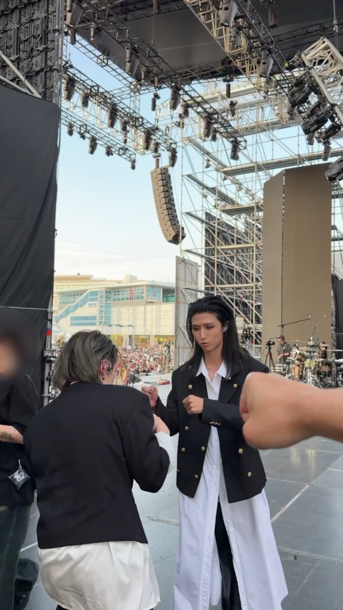 A performer in a black and white blazer outfit stands on stage with a long-haired drummer ("Guitarz") who has turned away from his kit, appearing to confront or punch him during a concert, matching the chaotic post-concert moment described.