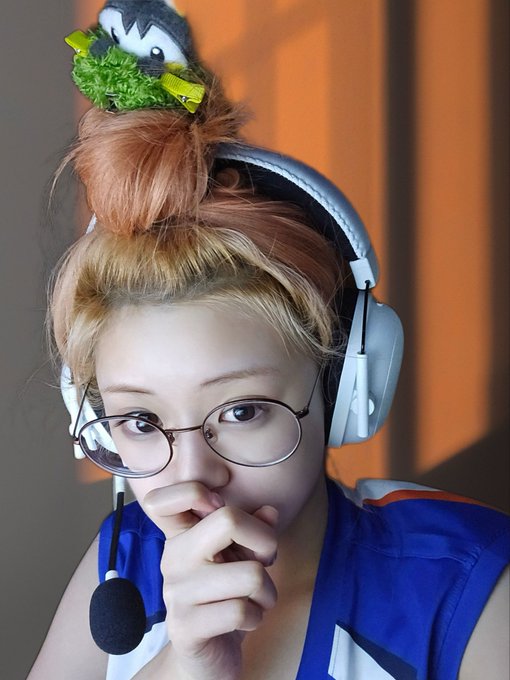 A young woman wearing a headset with a microphone and round glasses, with her hair in a bun topped by a small plush bird.