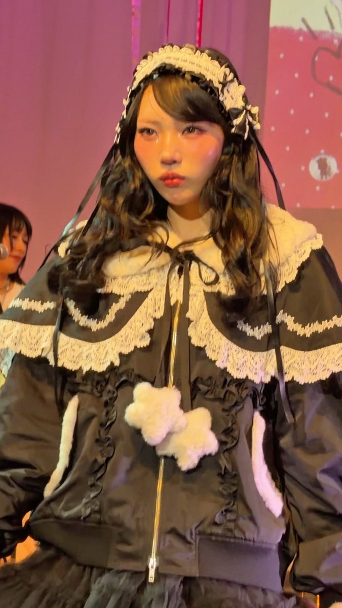 A young model with long dark hair poses confidently in a gothic lolita outfit, featuring a black hooded cape with cream lace trim, ribbon details, and a fluffy white cross appliqué on the chest. The image captures her at a Candy Stripper event, matching the post's mention of Miu-chan modeling the brand's signature Phantom Sita collection.