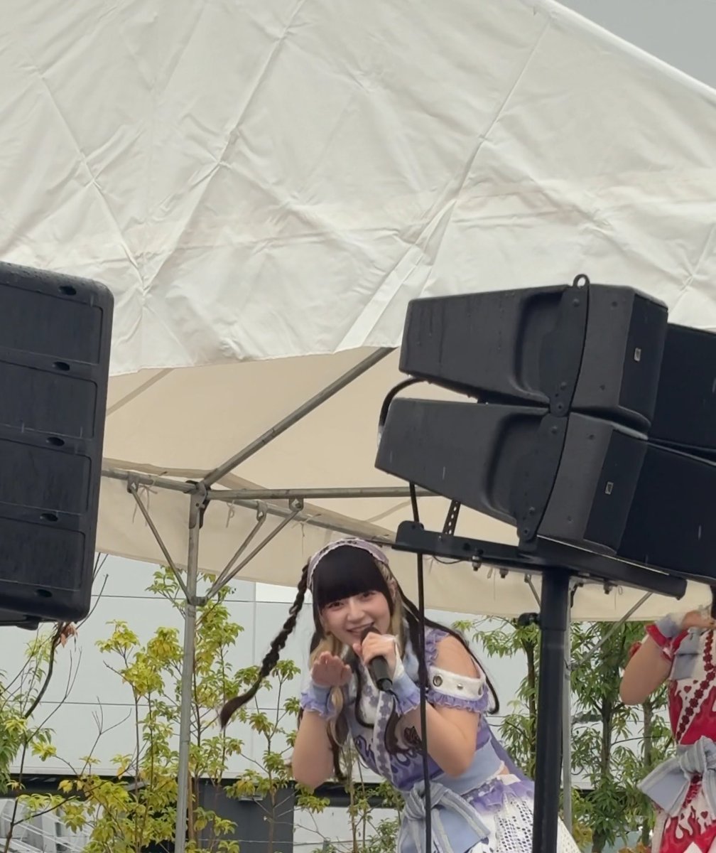 A performer in a purple and white frilly cosplay outfit speaks into a microphone on an outdoor stage beneath a tent canopy and rigging.