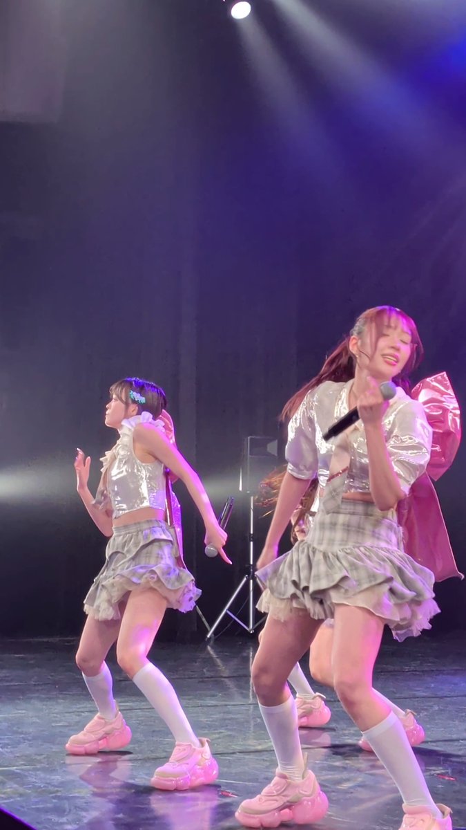 Two performers from the Japanese idol group onefive (members SOYO, KANO, GUMI, and MOMO) energetically dance on stage at the 10/3 Kanazawa Kagayaki Festival, wearing coordinated silver crop tops, ruffled skirts, and white knee-high socks under purple stage lighting.