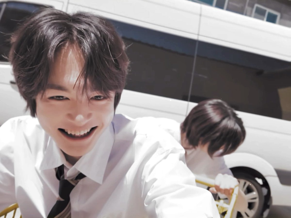 A young man with brown hair wearing a white shirt and tie smiles excitedly at the camera, with another person in similar attire visible in the background near a vehicle.