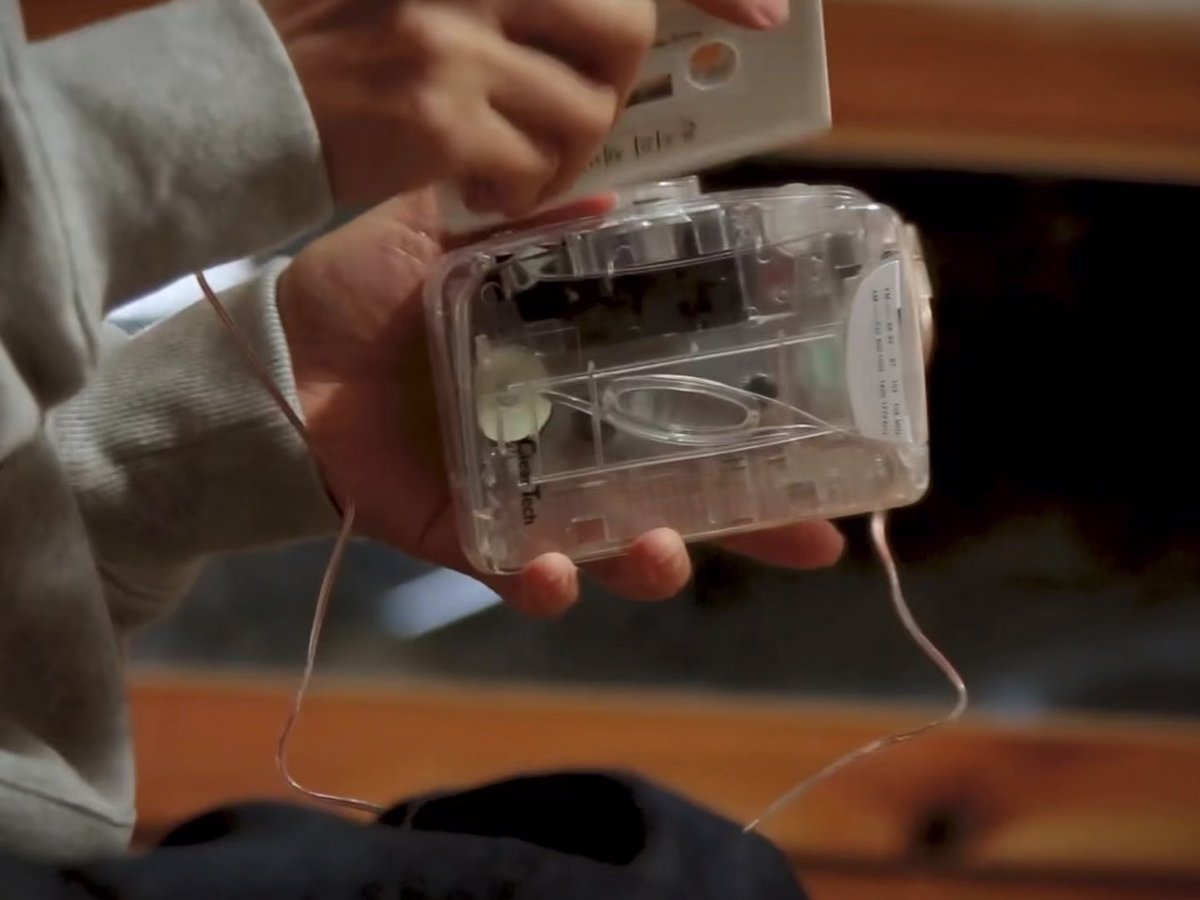 A close-up photo of a person's hands holding the internal mechanical components of a small clear plastic cassette tape player, with the front cover being lifted off, showing the tape reels and electronics.