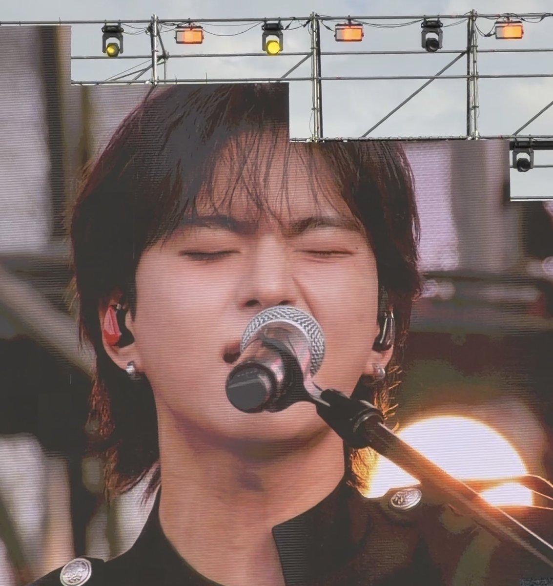 A close-up of a person singing into a microphone with their eyes closed, performing on a stage with overhead lighting rigs and a large screen visible in the background.