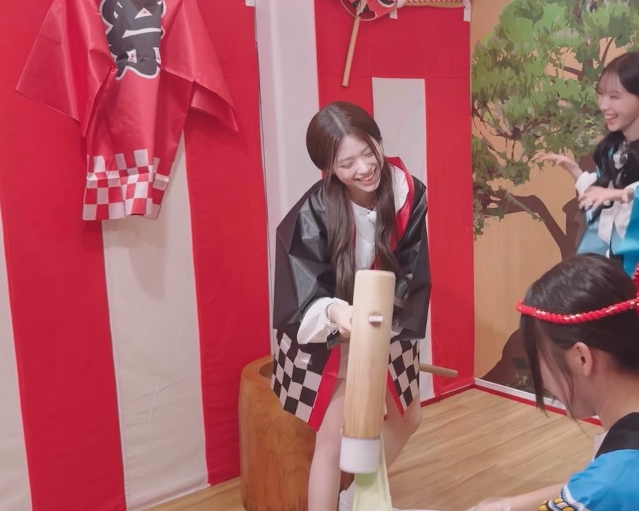 A smiling young woman in a black and white checkered kimono with red accents swings a wooden mallet at a traditional Japanese mochitsuki (rice cake) pounding station, with another woman watching in the background.