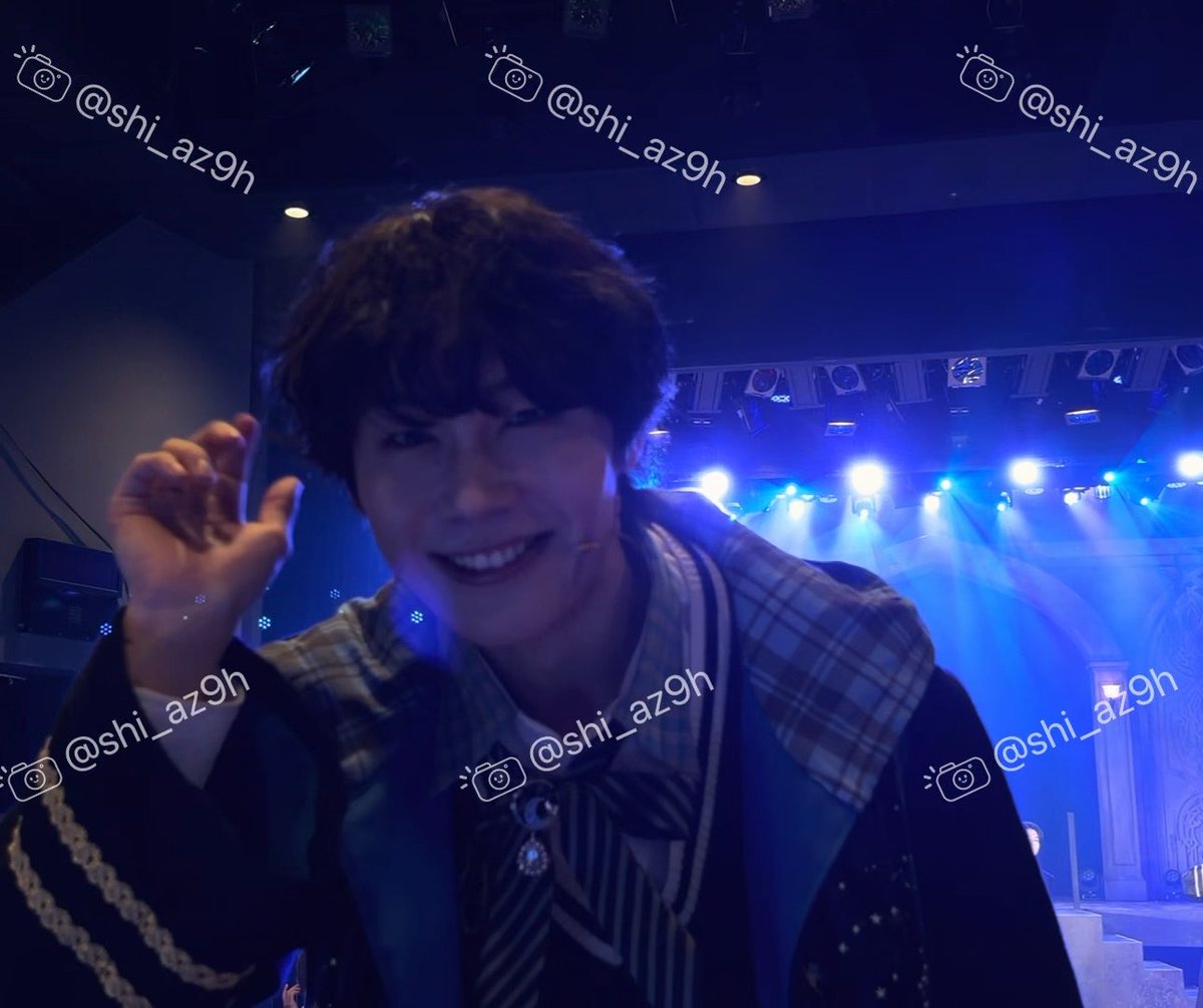 A person in a Japanese idol-style school uniform costume posing and smiling at the camera on a stage lit with blue lights, watermarked with @shi_az9h.