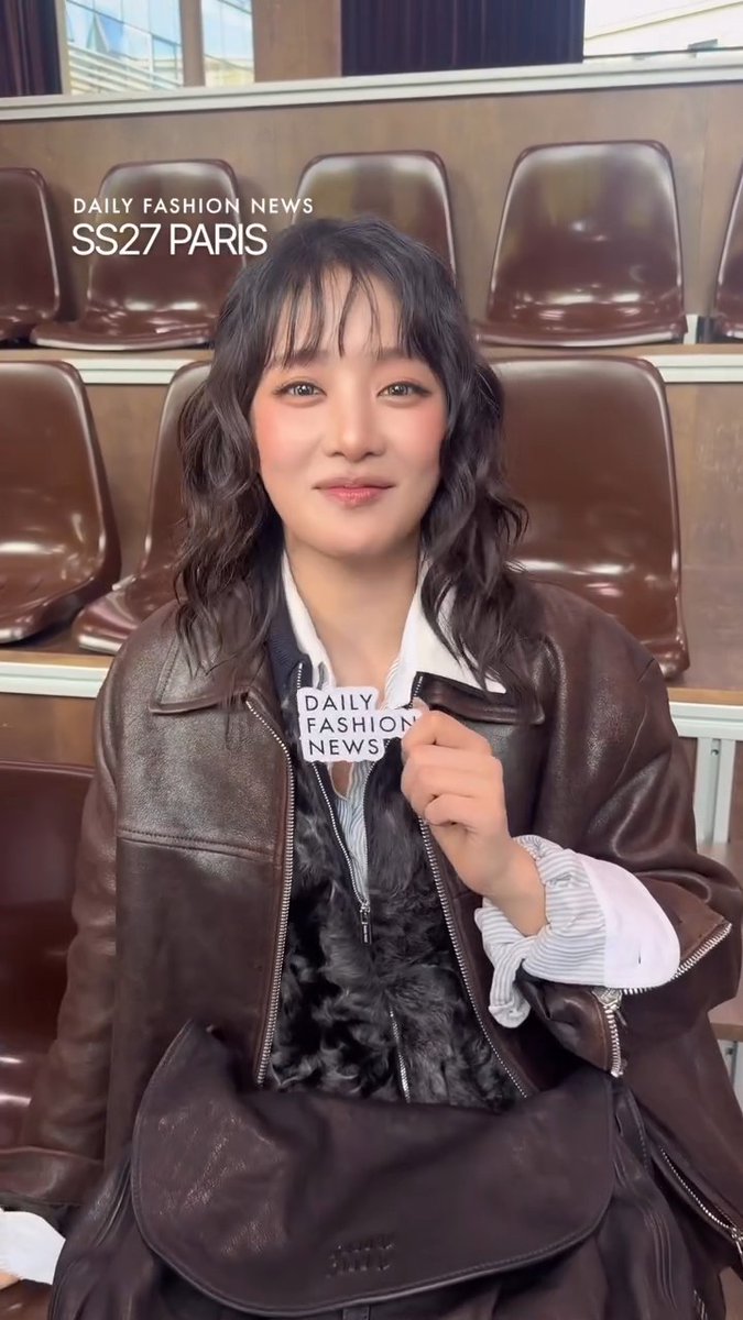 A woman with wavy dark hair sits in brown leather stadium-style seats, wearing a brown leather jacket and holding a "DAILY FASHION NEWS" microphone, with text overlay reading "DAILY FASHION NEWS SS27 PARIS."