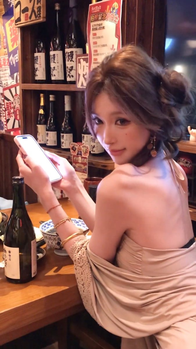 A woman in a beige backless dress sits at a wooden bar in what appears to be a Japanese restaurant, holding up a smartphone with a white screen while looking over her shoulder; shelves behind her are lined with sake bottles and Japanese menu signs.