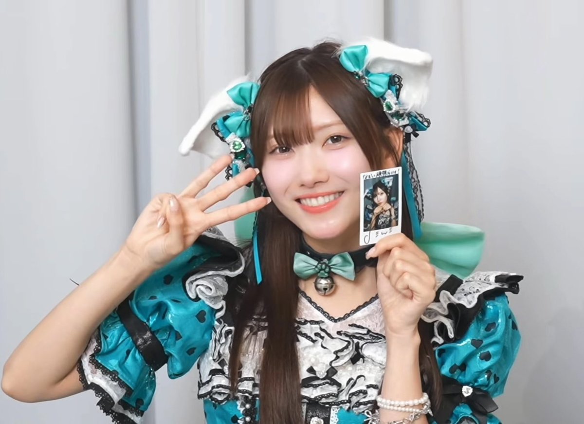 A smiling young woman wearing a teal and white frilly outfit with a small bell collar, posing with one hand up and holding a small polaroid photo in the other.