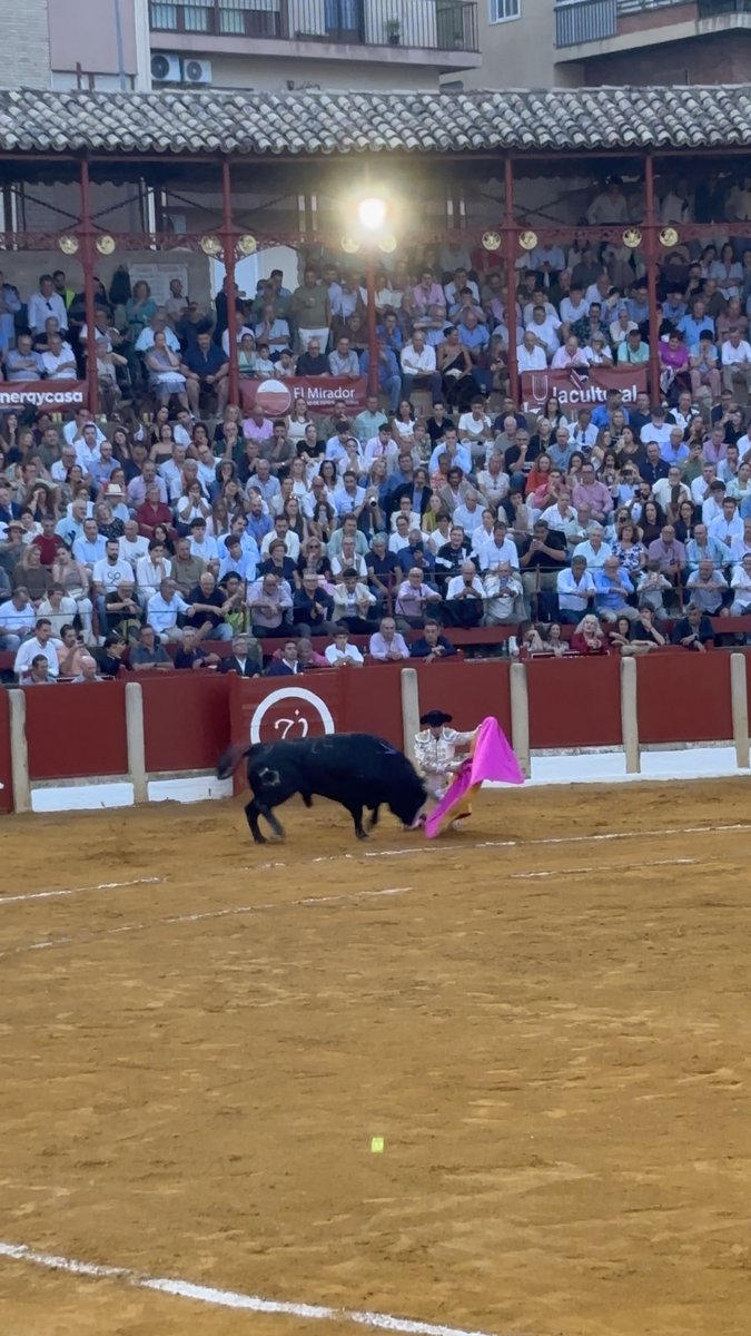 A matador in traditional attire uses a pink cape to engage a black bull in a dirt arena, with a crowded grandstand and signage reading 'El Mirador' and 'U Cultural' in the background.