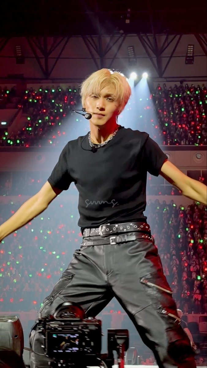 NCT 127's Haechan, sporting blonde hair and a black t-shirt with double studded belts, performs energetically on stage during the Neo City – The Redline tour stop in Jakarta, with fans waving colorful lightsticks behind him.